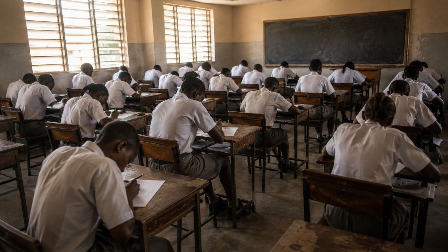 A full secondary school classroom during an examination: rows of students in uniform at wooden desks, heads down, writing on answer sheets in daylight from louvred windows.