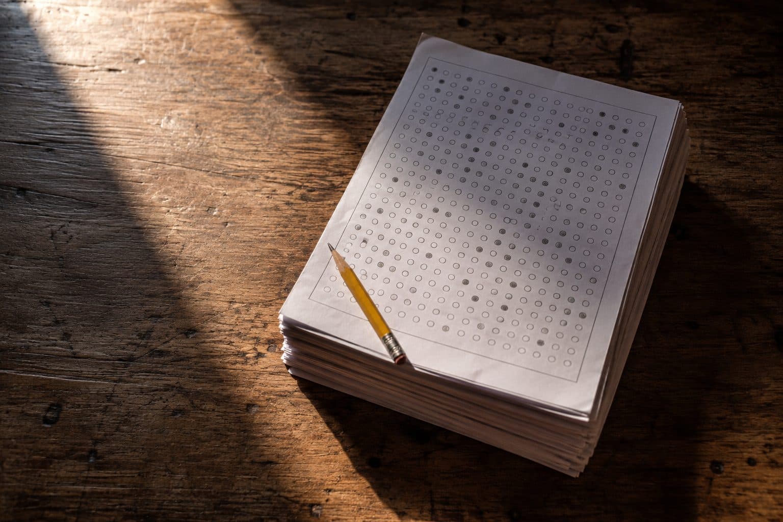 A squared stack of completed multiple-choice answer sheets on a worn wooden desk, a yellow pencil resting across the top sheet in low afternoon light.